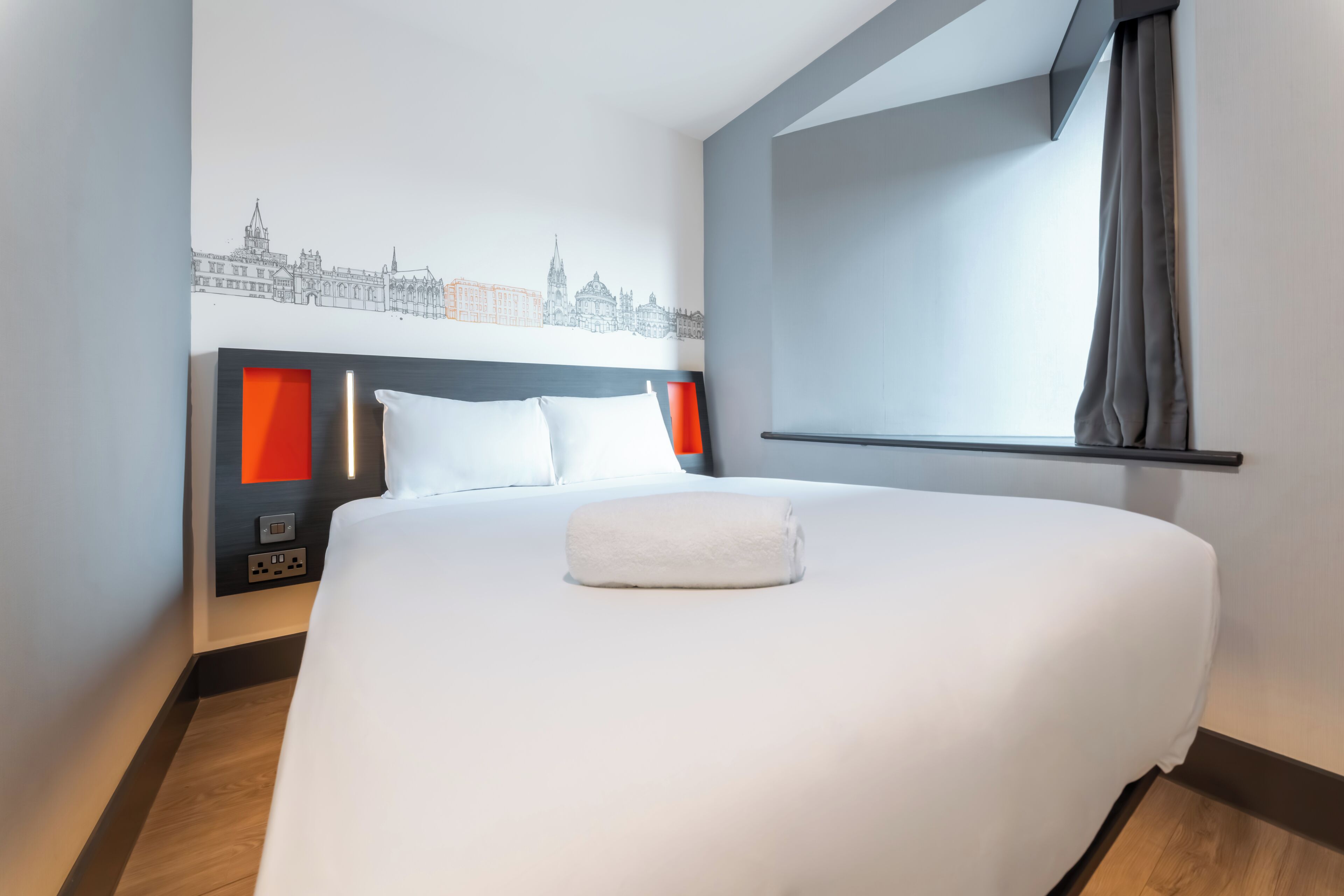 A hotel room with a large bed, a cityscape mural on the wall, a window with curtains, and a bedside table with a lamp.