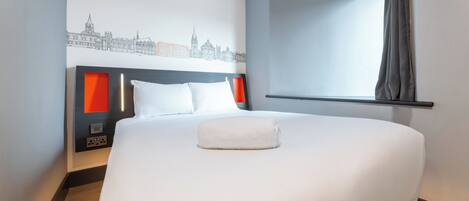 A hotel room with a large bed, a cityscape mural on the wall, a window with curtains, and a bedside table with a lamp.