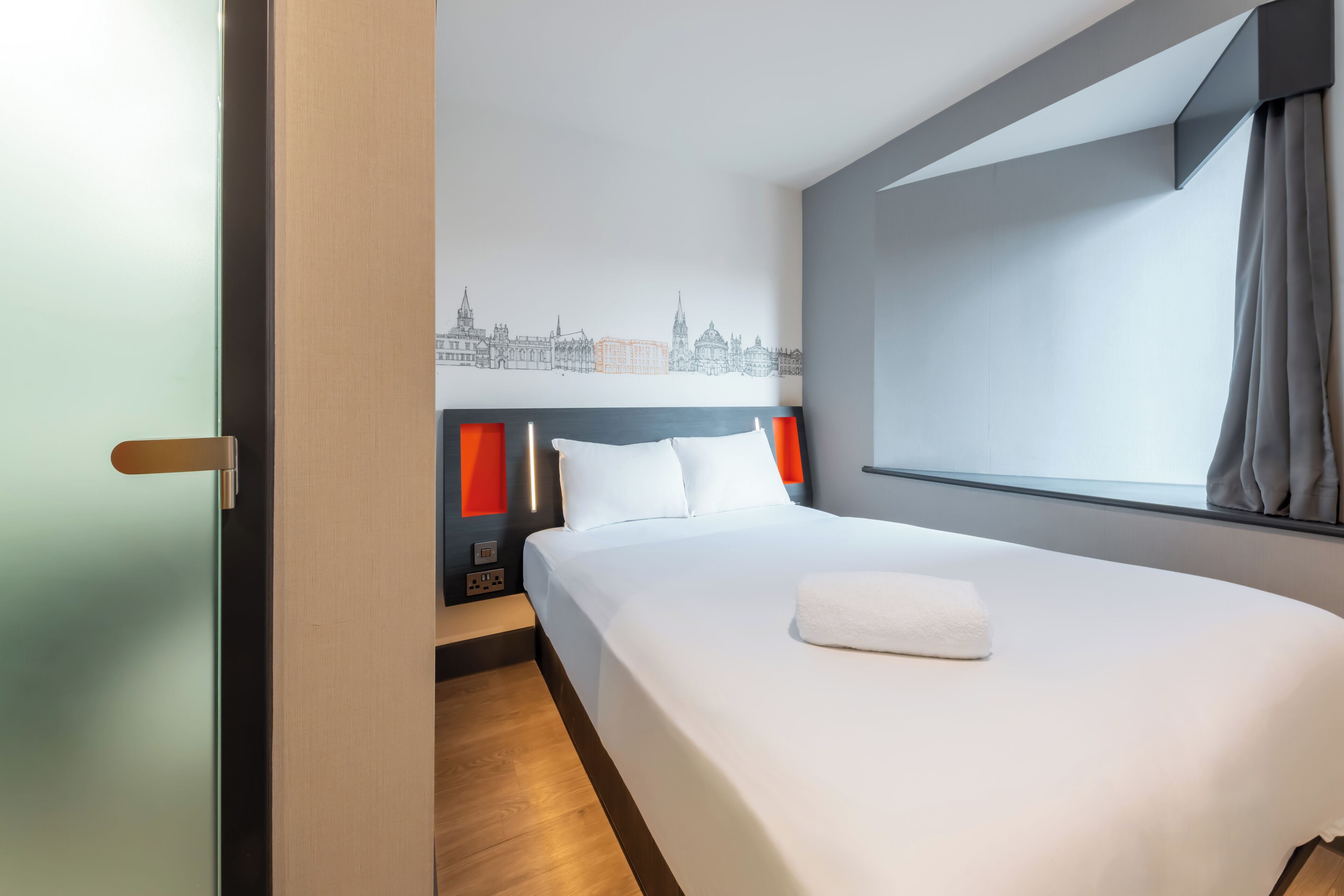 Una habitación de hotel con una cama, un mural de paisaje urbano en la pared, una ventana con cortinas y suelo de madera.