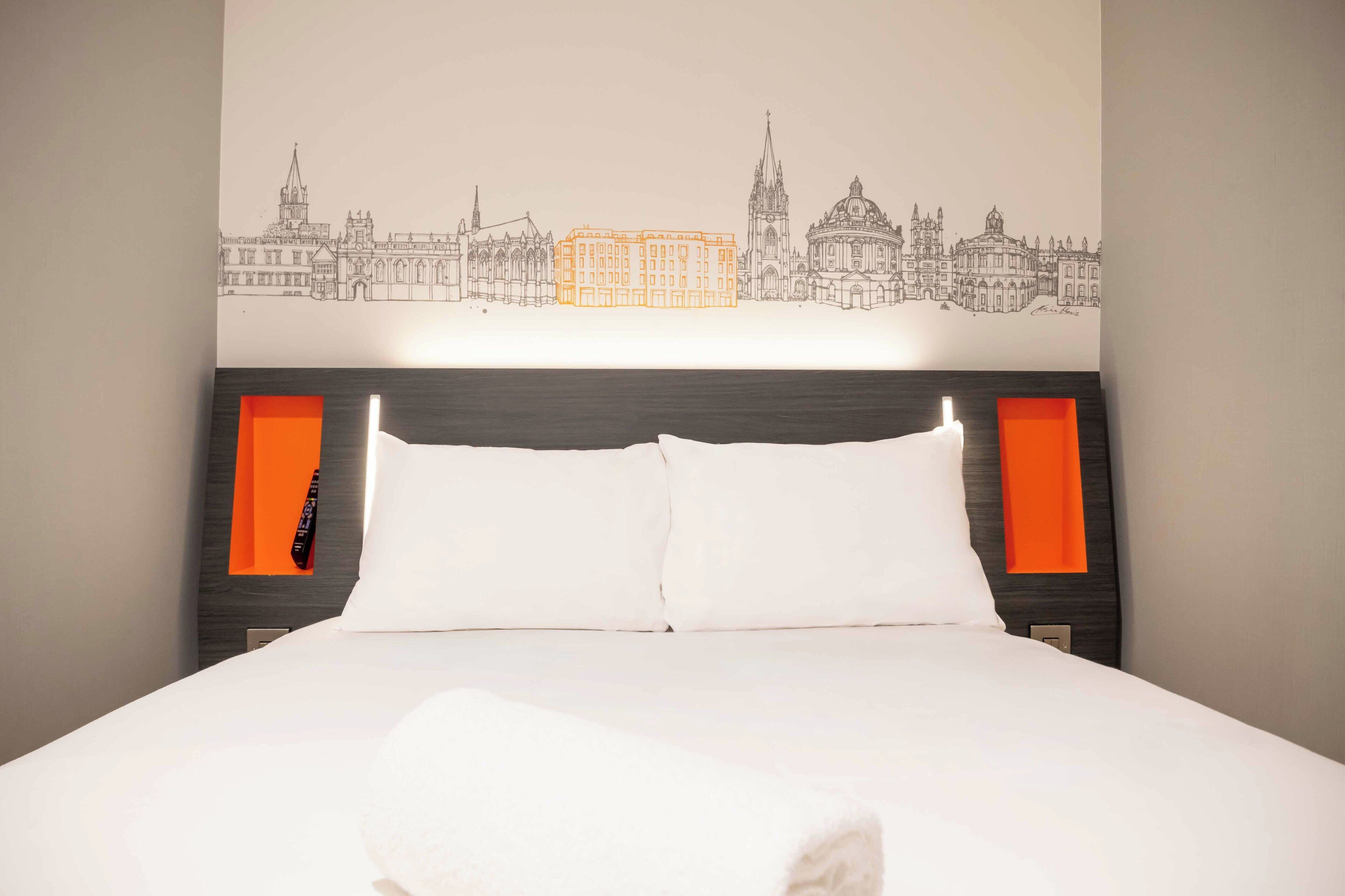 A hotel room with a bed, white bedding, a cityscape wall art, and orange accents on the headboard and nightstand.
