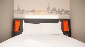 A hotel room with a bed, white bedding, a cityscape wall art, and orange accents on the headboard and nightstand.