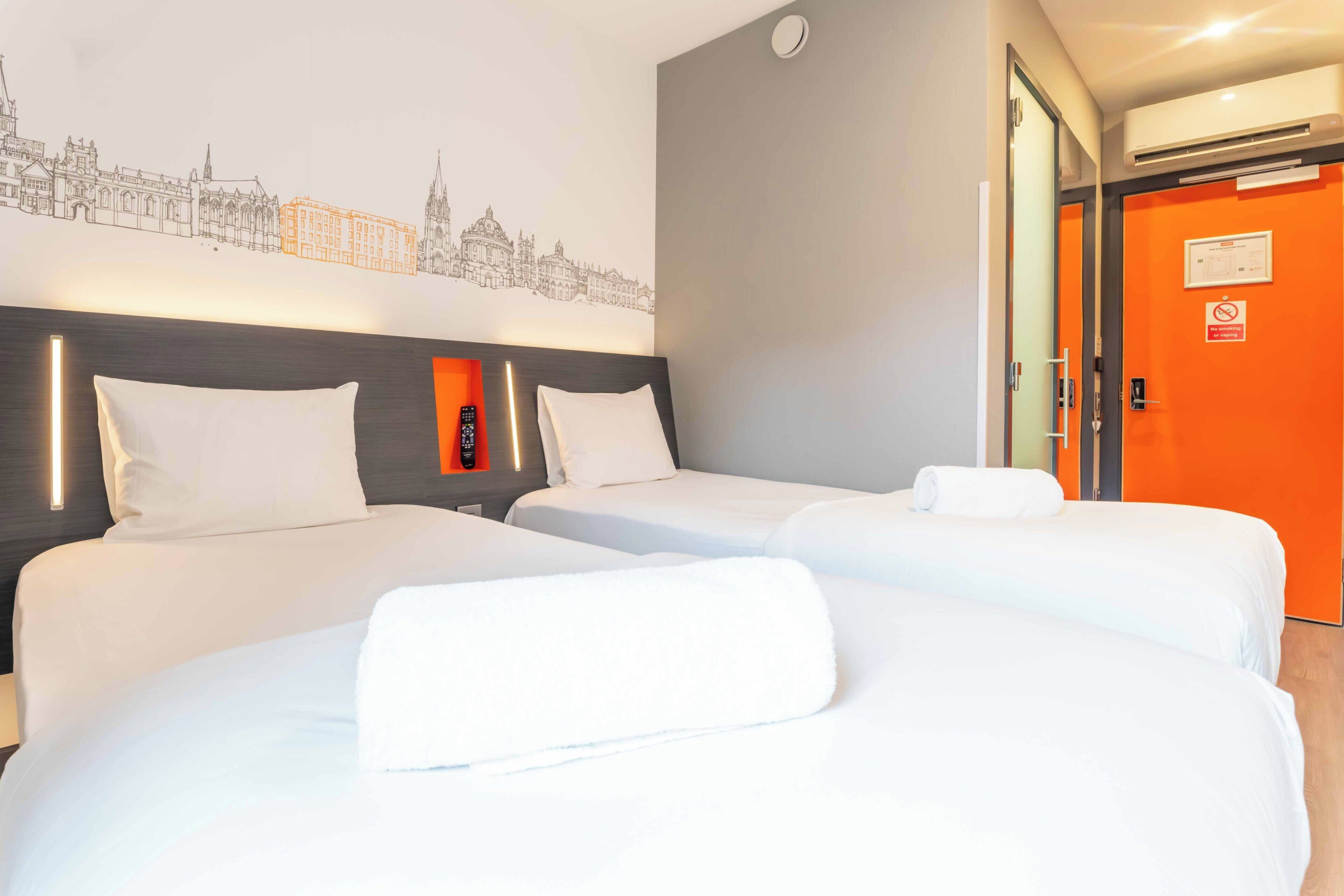 Une chambre d’hôtel avec deux lits, une décoration murale représentant un paysage urbain, une tête de lit moderne et une porte d’un orange vif.