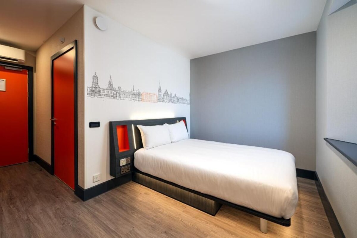 A hotel room with a bed, a red door, and a cityscape mural on the wall.