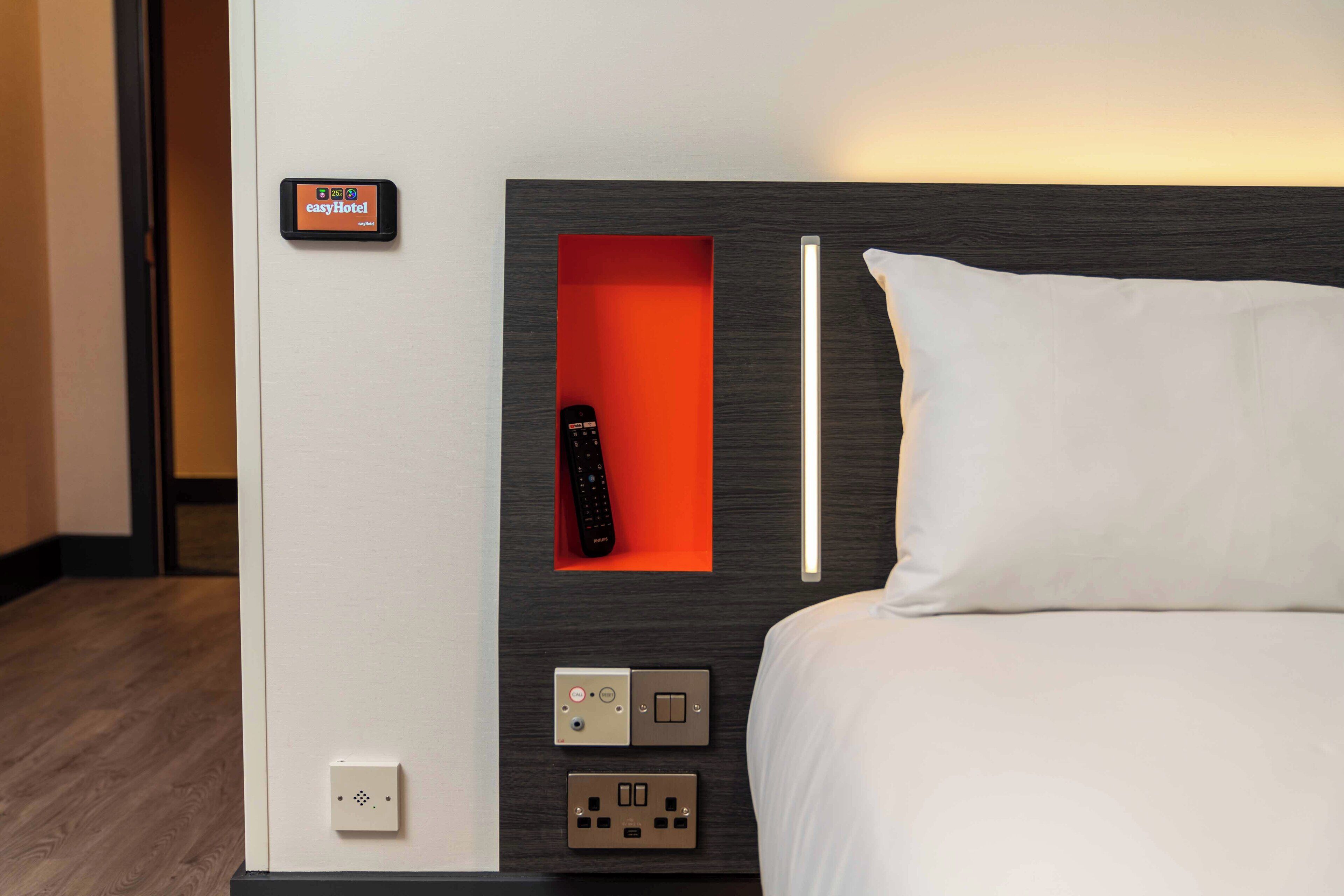 Une chambre d’hôtel avec un lit, une télécommande dans un compartiment rouge et un panneau de commande fixé au mur, doté de nombreux boutons et interrupteurs.