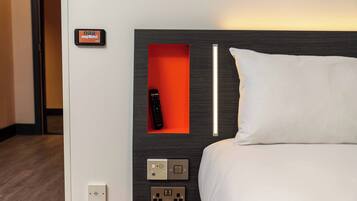 A hotel room with a bed, a remote control in a red slot, and a wall-mounted control panel with multiple buttons and switches.