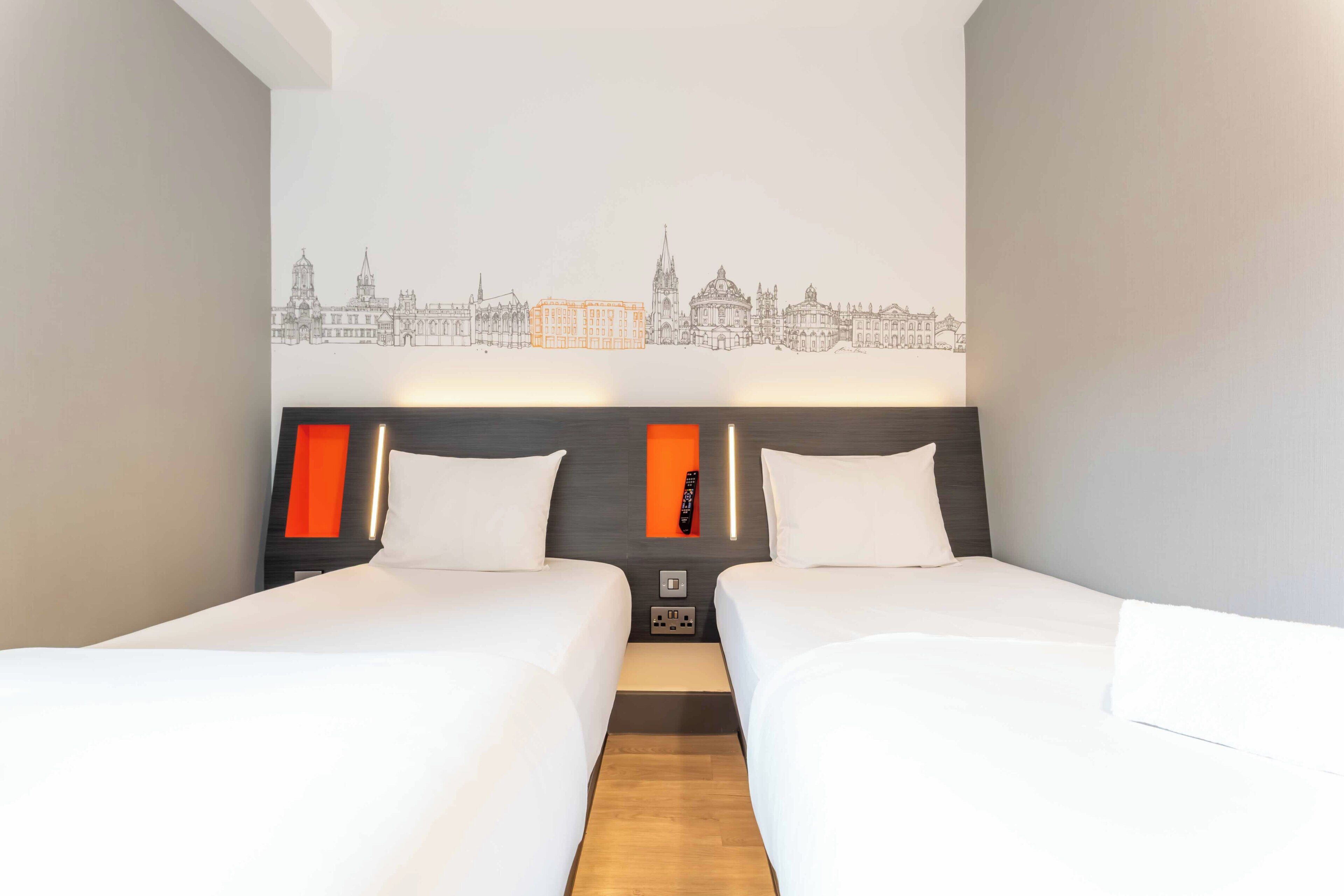 A hotel room with two beds, a cityscape wall art, and a modern headboard with orange accents.