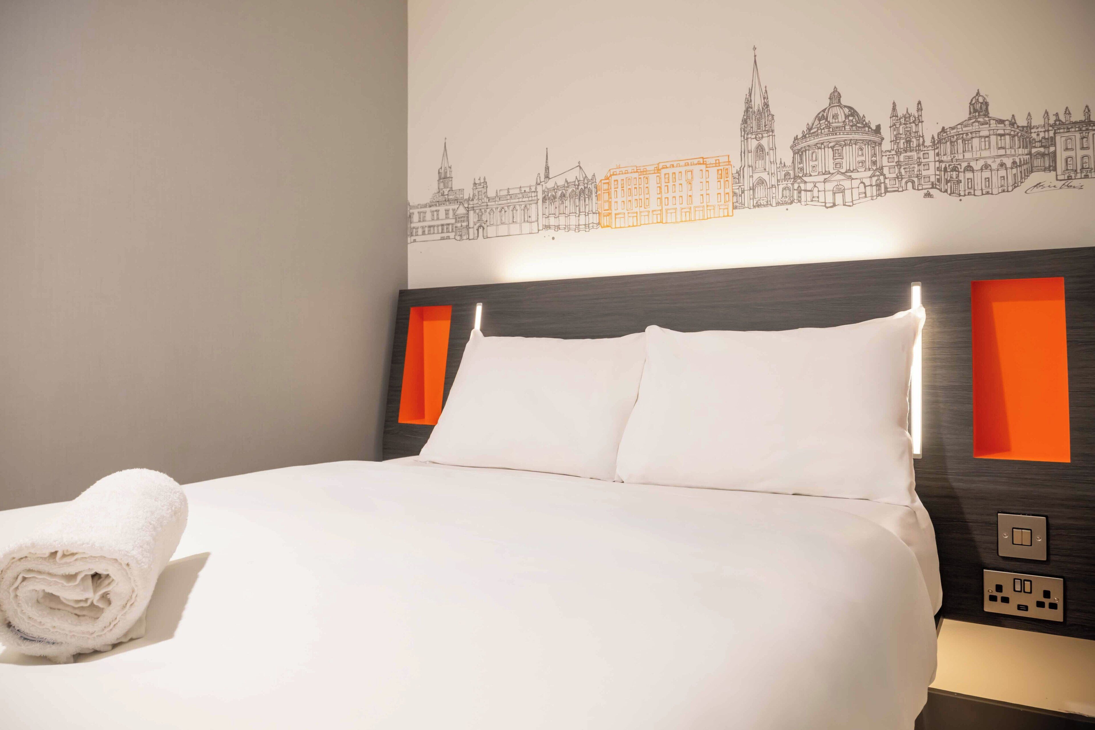 A hotel room with a neatly made bed, a cityscape mural on the wall, and a modern headboard with integrated lighting.
