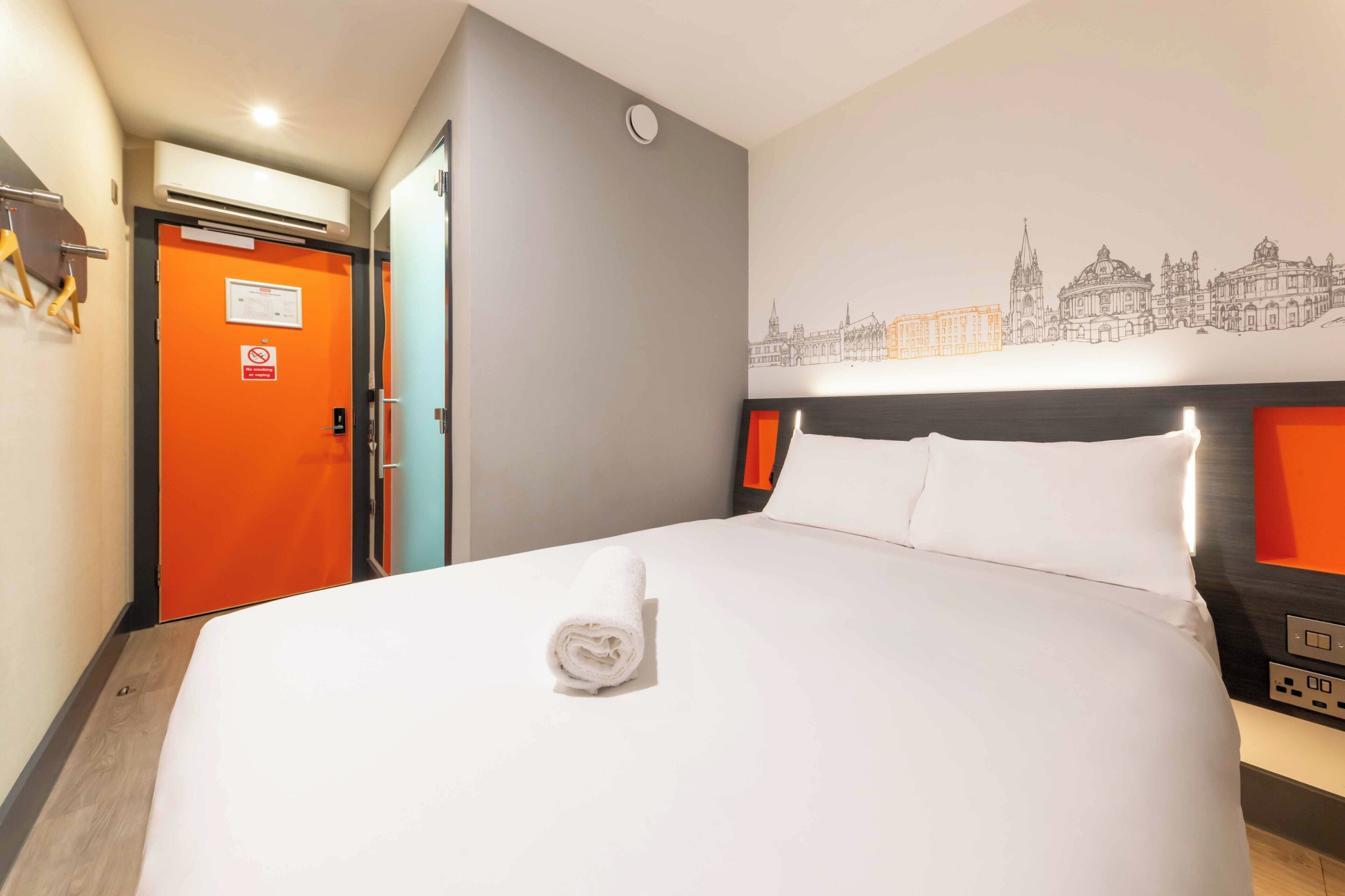 Een hotelkamer met een net opgemaakt bed, stadsbeeld wandkunst, een oranje deur en een bedieningspaneel aan de zijkant van het bed.