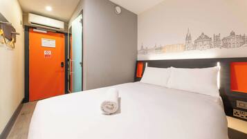 A hotel room with a neatly made bed, a cityscape wall art, an orange door, and a control panel on the bed's side.