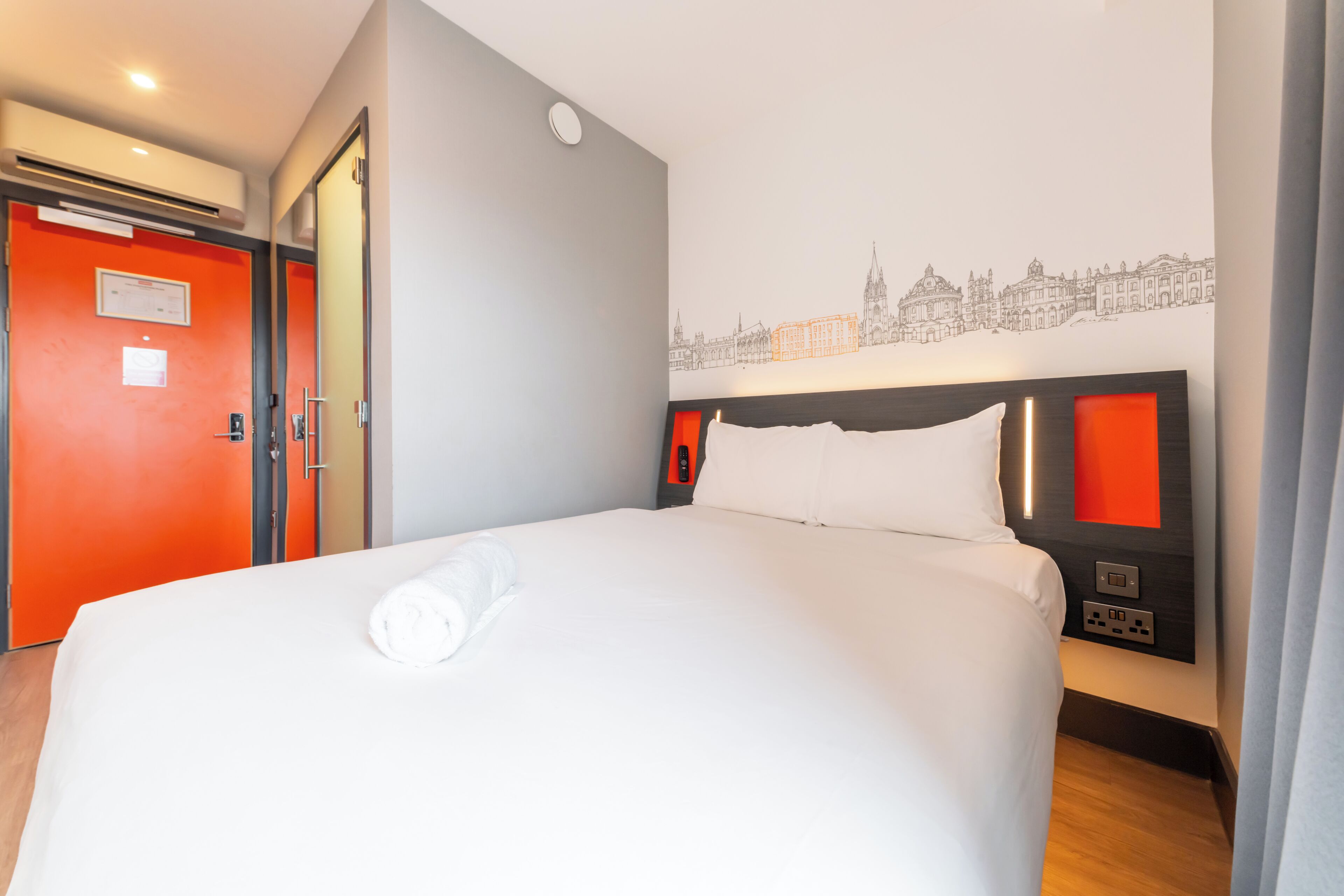 A hotel room with a bed, a red door, a cityscape wall art, and a headboard with built-in lighting.