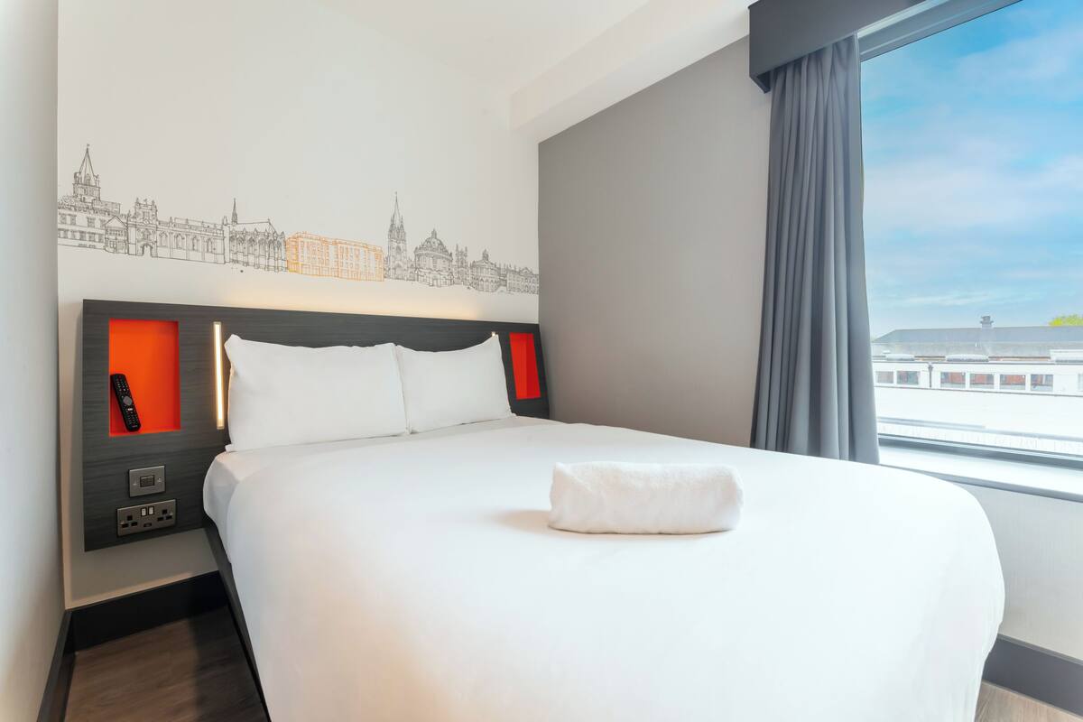 Une chambre d’hôtel avec un grand lit, une décoration murale représentant un paysage urbain, une fenêtre avec vue et une table de chevet avec une télécommande.