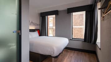 Quarto de hotel moderno com cama, janela com cortinas, parede de tijolos visível pela janela e piso de madeira.