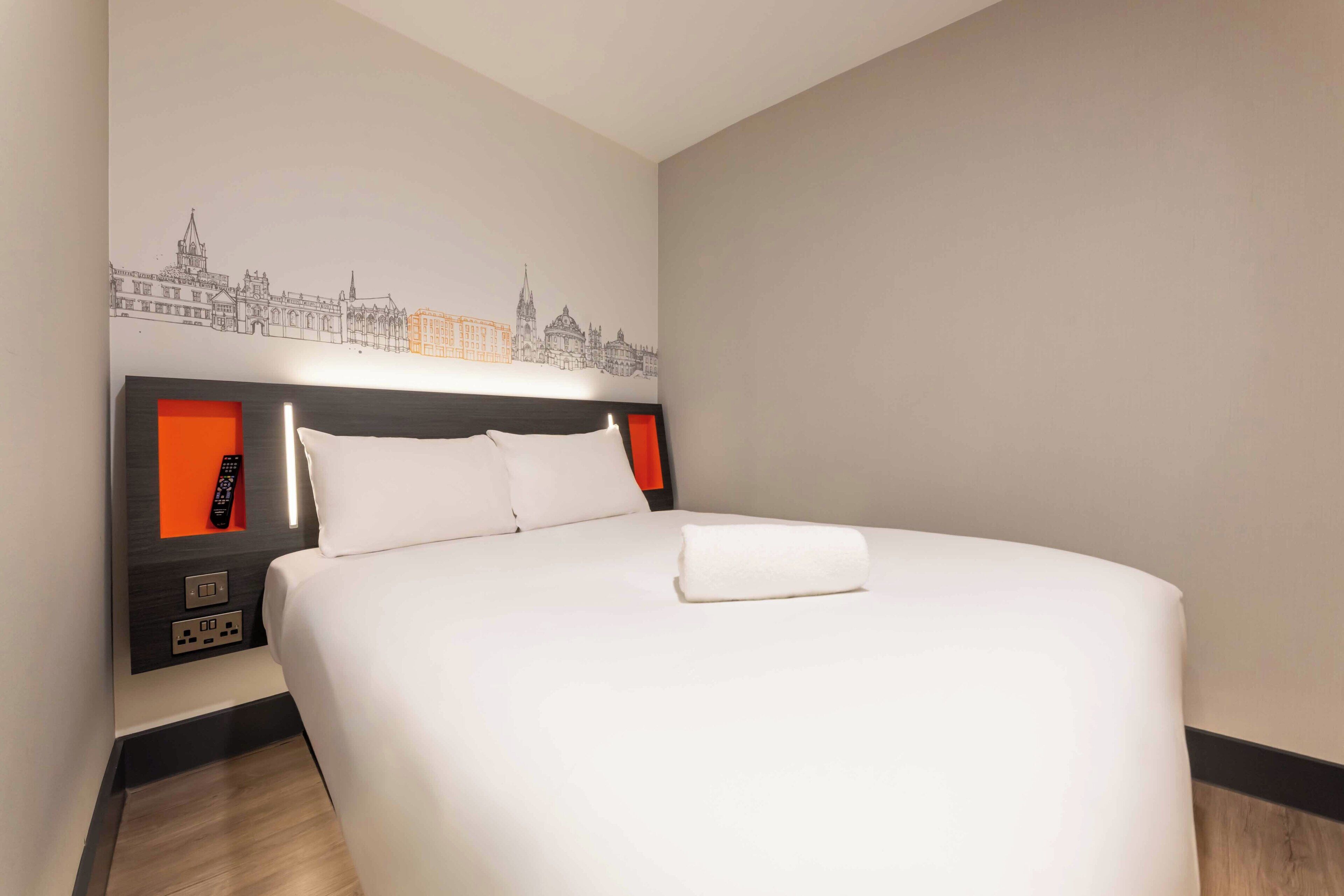 Habitación de hotel con una cama grande, cabecera con control remoto y un cuadro en la pared que representa un paisaje urbano.