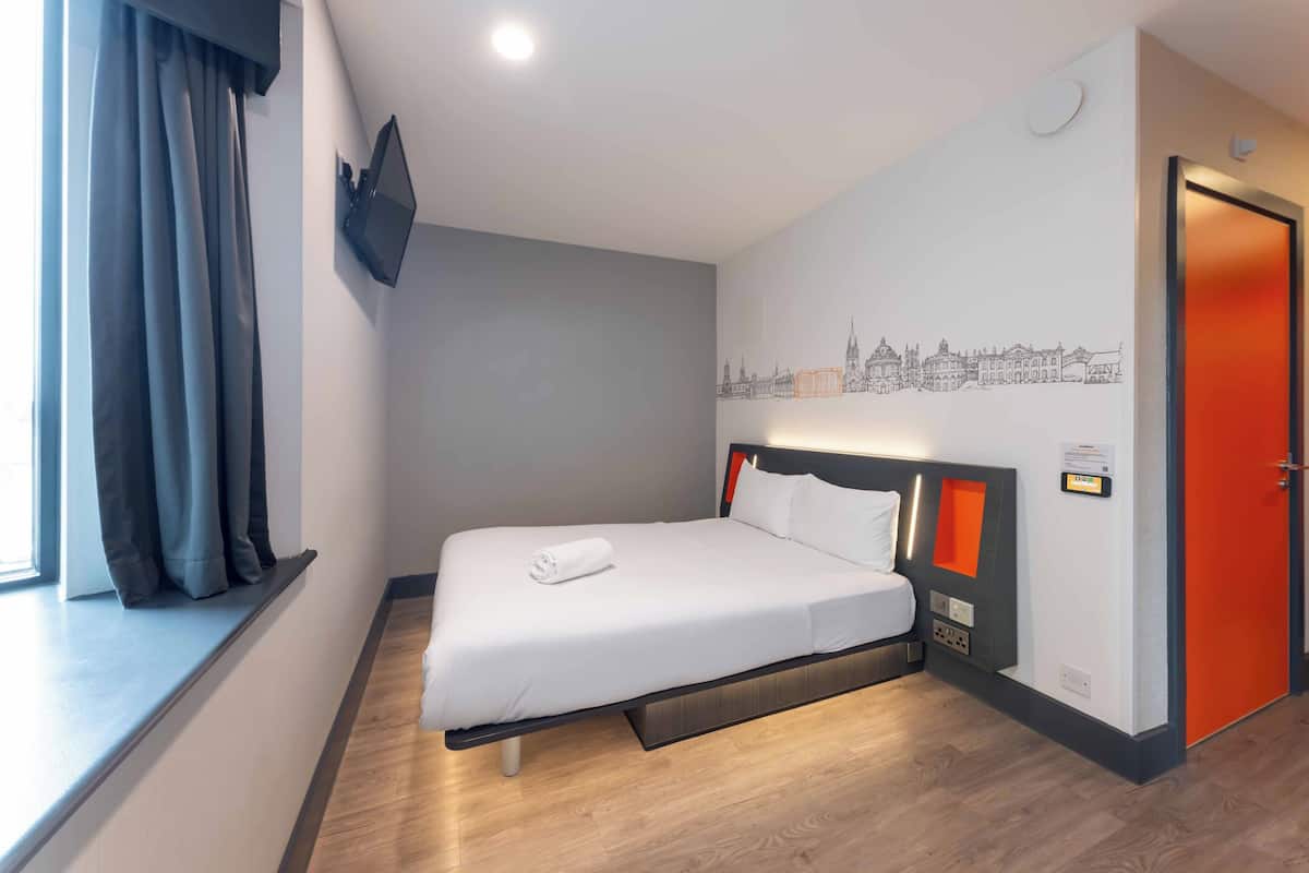 Une chambre d’hôtel avec un lit, une fenêtre avec des rideaux, un téléviseur fixé au mur et une porte.