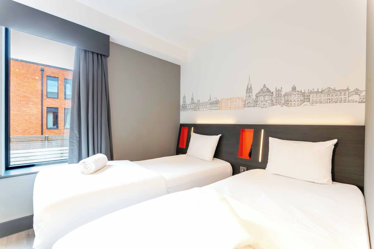 Une chambre d’hôtel avec deux lits, une fresque représentant un paysage urbain, une fenêtre avec des rideaux et une vue sur un immeuble en briques.