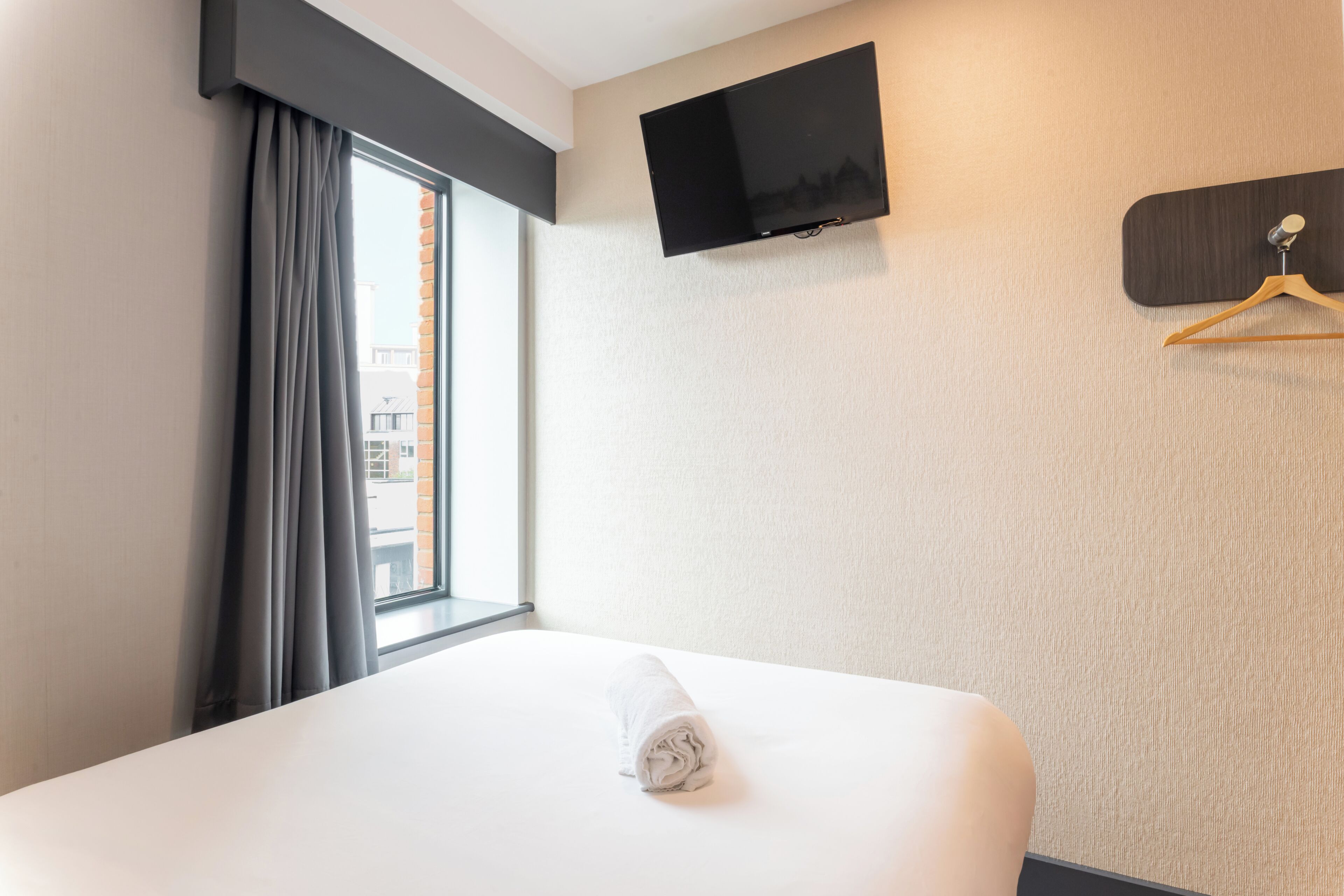 Ein Hotelzimmer mit Bett, einem an der Wand befestigten Fernseher, einem Fenster mit Vorhängen und einem hölzernen Kleiderhaken an der Wand.