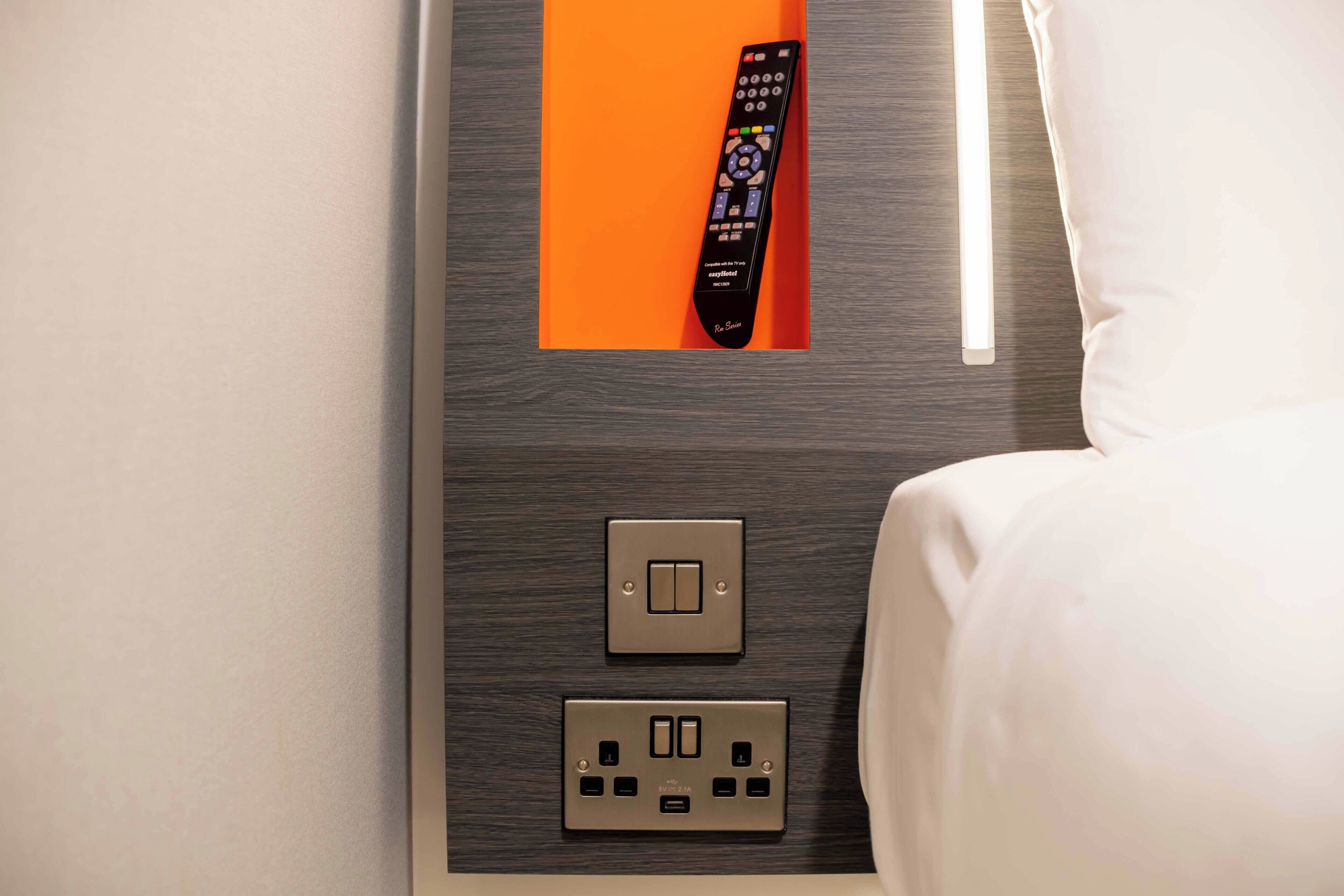 A hotel room with a bed, a remote control, a light switch, and a power outlet.
