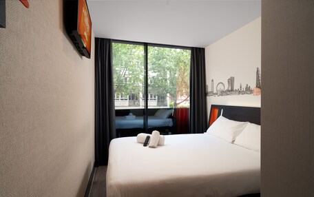 A hotel room with a large bed, a flat-screen TV mounted on the wall, and a window with curtains.