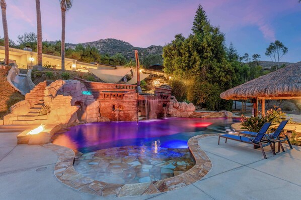 A swimming pool with a waterfall feature, surrounded by stone architecture and palm trees.
