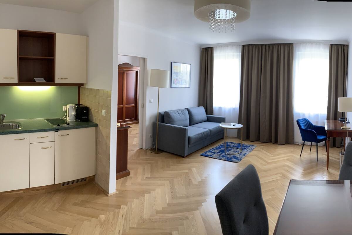 A modern hotel room with a kitchenette, a sofa, a dining table, and a TV.