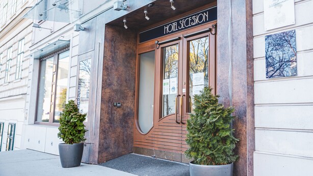A hotel entrance with a wooden door, two potted plants, and a sign indicating it is the 5th building on the street. - Hotel Secession an der Oper (Vienna)