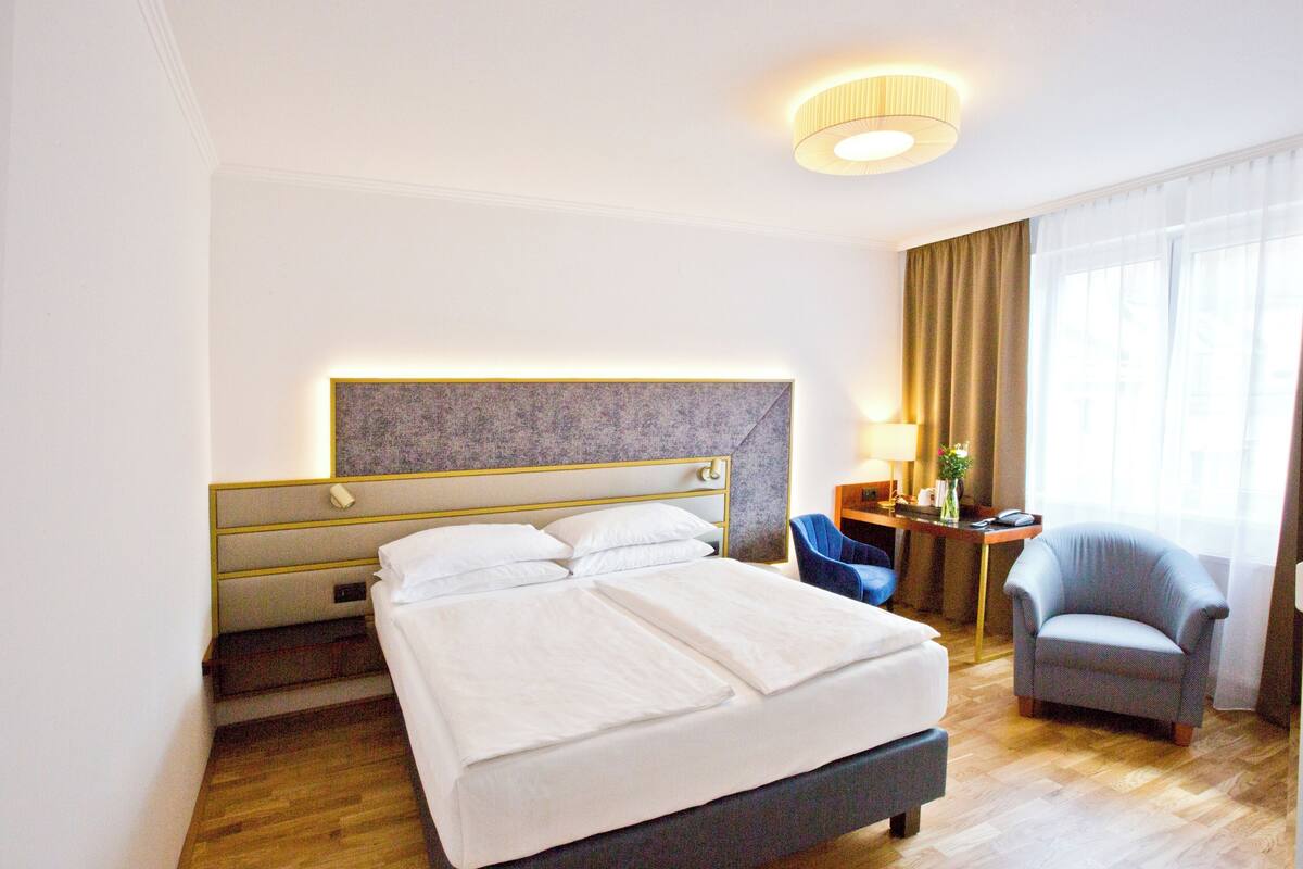 A hotel room with a bed, a desk, a chair, and a large window with curtains.