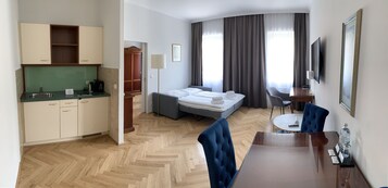 Una camera d'albergo con angolo cottura, un letto con testiera grigia, un tavolo da pranzo con sedie, una televisione e un'ampia finestra con tende.