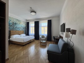 Quarto de hotel com cama, poltrona, escrivaninha e televisão.