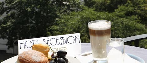 Una mesa con un cartel que dice "HOTEL SECESSION", un plato de comida y una jarra de cerveza, con vistas a un paisaje urbano que incluye edificios y una cúpula.
