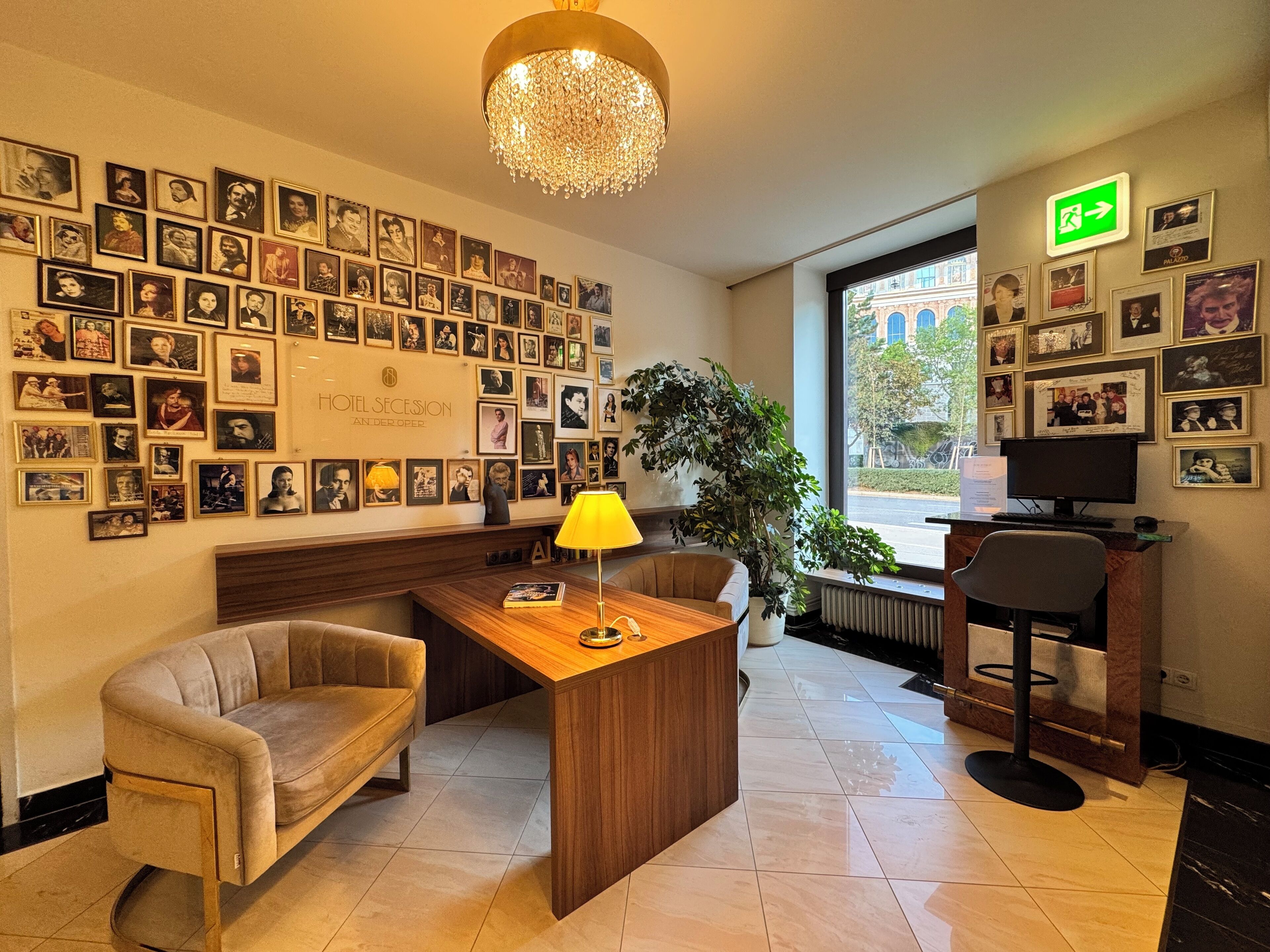 A hotel lobby with a wall of framed photos, a wooden reception desk, a yellow lamp, a potted plant, and a large window with a view of buildings outside.