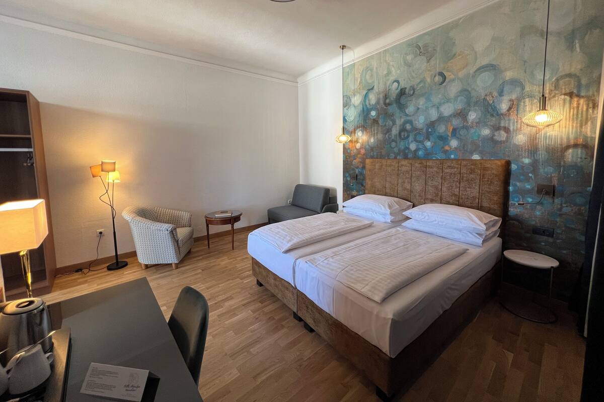 A hotel room with a large bed, a desk with a chair, a small table, a chair, and a wall with a blue abstract design.