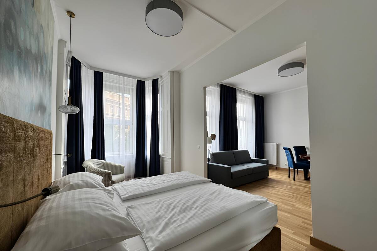 A modern hotel room with a large bed, a sofa, a dining area, and a view of the city.