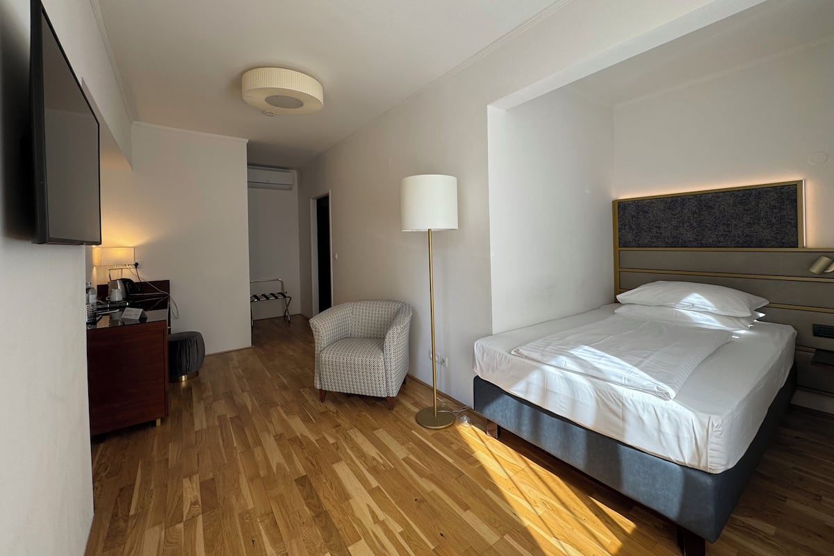 A hotel room with a bed, a chair, a desk with a lamp, a flat-screen TV, and a ceiling light.
