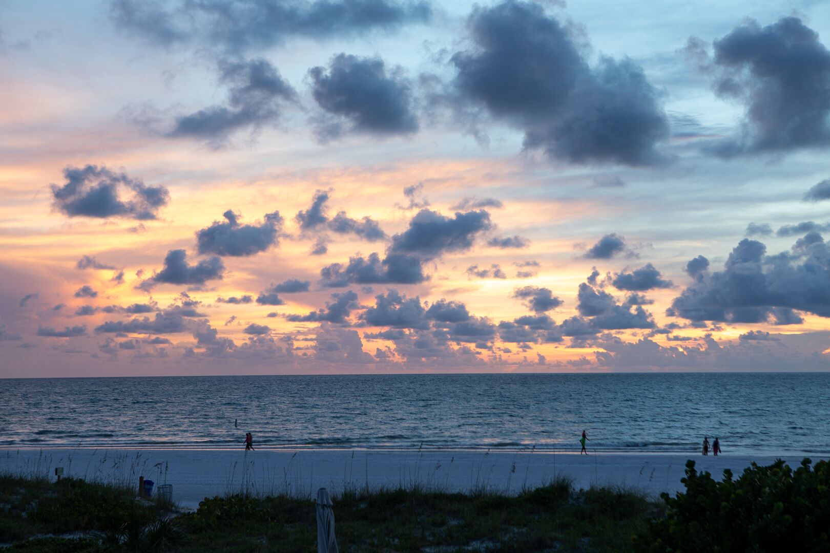 A beach with people walking along the shore, a clear sky with clouds, and the ocean in the background.