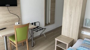A hotel room with a wooden desk, a green chair, a small table, a bed with patterned bedding, a TV mounted on the wall, and a mirror. - Gasthof Linde-Classico (Bad Mergentheim)