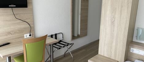 Habitación de hotel con un escritorio de madera, una silla verde, una mesita pequeña, una cama con ropa de cama estampada, un televisor montado en la pared y un espejo.