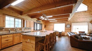A spacious kitchen with wooden cabinets, a large island with bar stools, a dining area with a sofa, and a clock on the wall. - Cozy Cabin with Hot Tub Near Yellowstone (Island Park)
