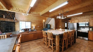 A spacious kitchen with wooden cabinets, a large island with bar stools, a stone fireplace, and a staircase leading to an upper level. - Cozy Cabin with Hot Tub Near Yellowstone (Island Park)