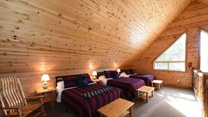 A cozy cabin with a sloped ceiling, wooden walls, and a large window. There are two beds with striped bedding and a rocking chair. - Cozy Cabin with Hot Tub Near Yellowstone (Island Park)