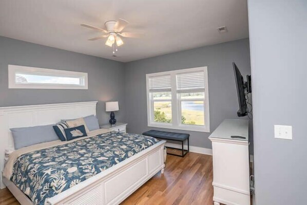A bedroom with a bed, a nightstand, a lamp, a bench, a ceiling fan, and a window with blinds. - A magical setting with ocean and marsh views! (Oak Island)