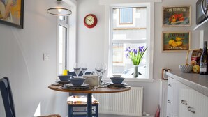 A dining area with a table set for four, a clock on the wall, and a window with a view of the outside. - Three Creeks Cottage (Portknockie)