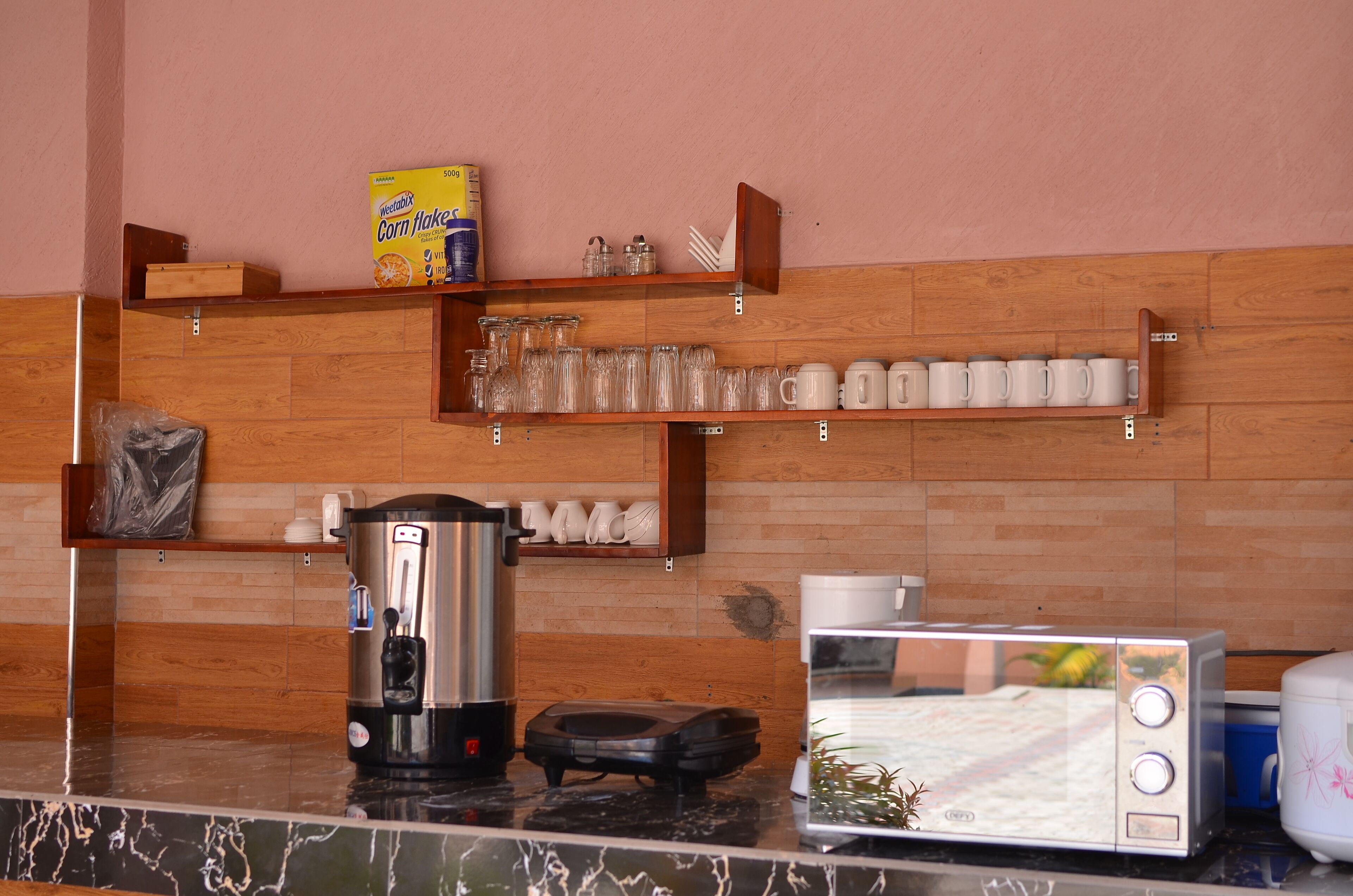 A kitchen with a coffee maker, toaster, and shelves with cups and a box of corn flakes.