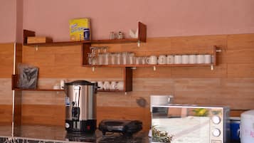 A kitchen with a coffee maker, toaster, and shelves with cups and a box of corn flakes.