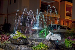 Uma fonte com jatos de água e uma estátua de cisne, rodeada por pedras e plantas.