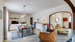 A modern kitchen with a marble countertop, dining area with chairs, and a wicker basket with a bottle and fruits.
