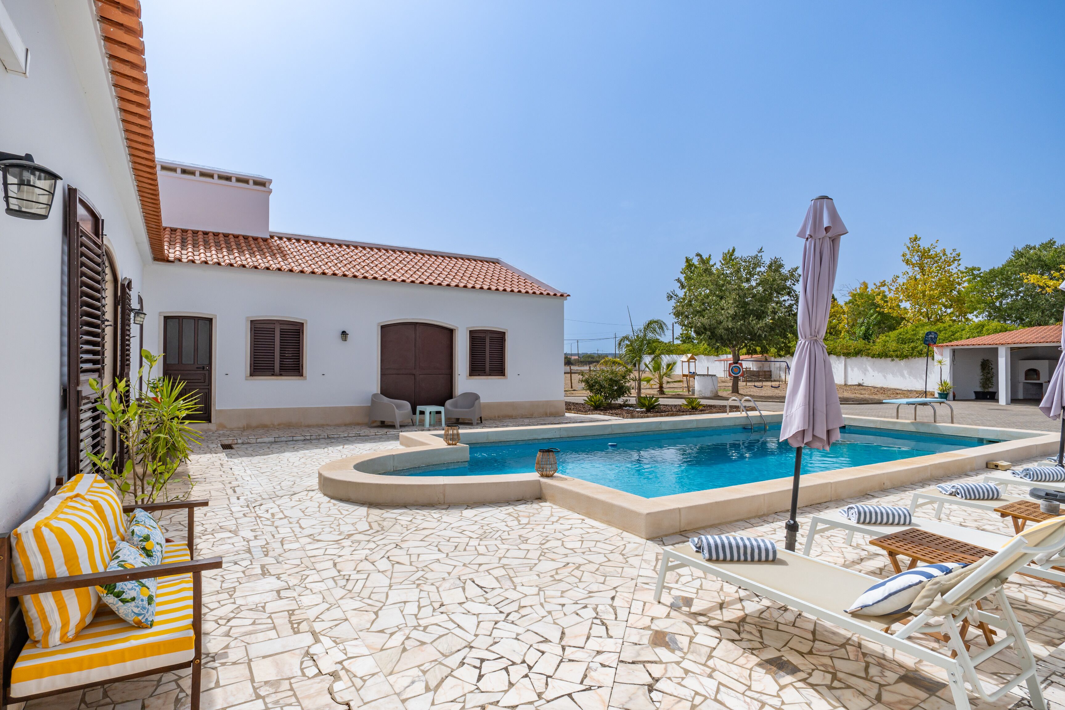 A pool area with lounge chairs and umbrellas, a tiled patio, and a building with a tiled roof.