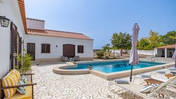 A pool area with lounge chairs and umbrellas, a tiled patio, and a building with a tiled roof.