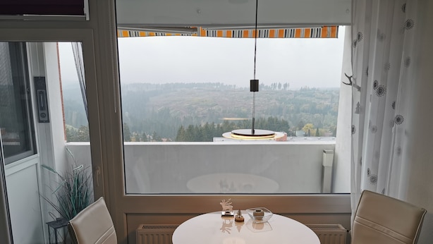 Ein Zimmer mit einem runden Tisch und zwei Stühlen, Blick auf eine Stadtlandschaft durch ein großes Fenster und ein Heizkörper unter dem Fenster.