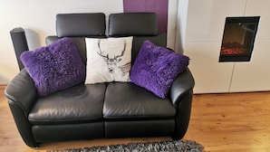 Ein schwarzes Ledersofa mit violetten, flauschigen Kissen und einem dekorativen Kissen mit Hirschmotiv, vor einem weißen Schrank mit Kamin.