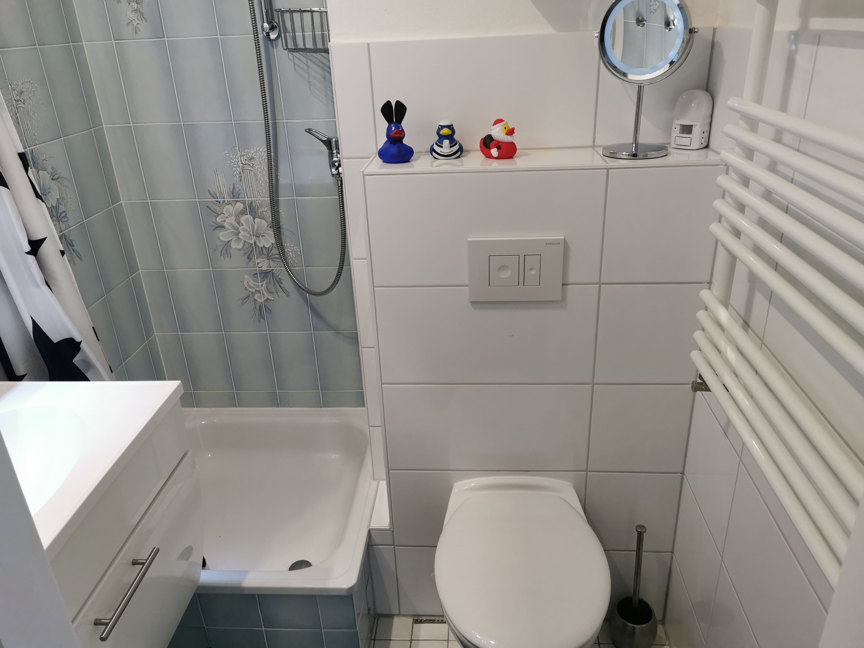 Une salle de bain équipée d’une baignoire, de toilettes et d’une douche. Les murs sont recouverts de carreaux décoratifs et une petite étagère contient des petits canards en caoutchouc.